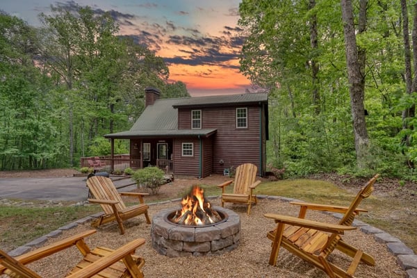 Red cabin surrounded by tall trees with stone fire pit and wooden Adirondack chairs at sunset